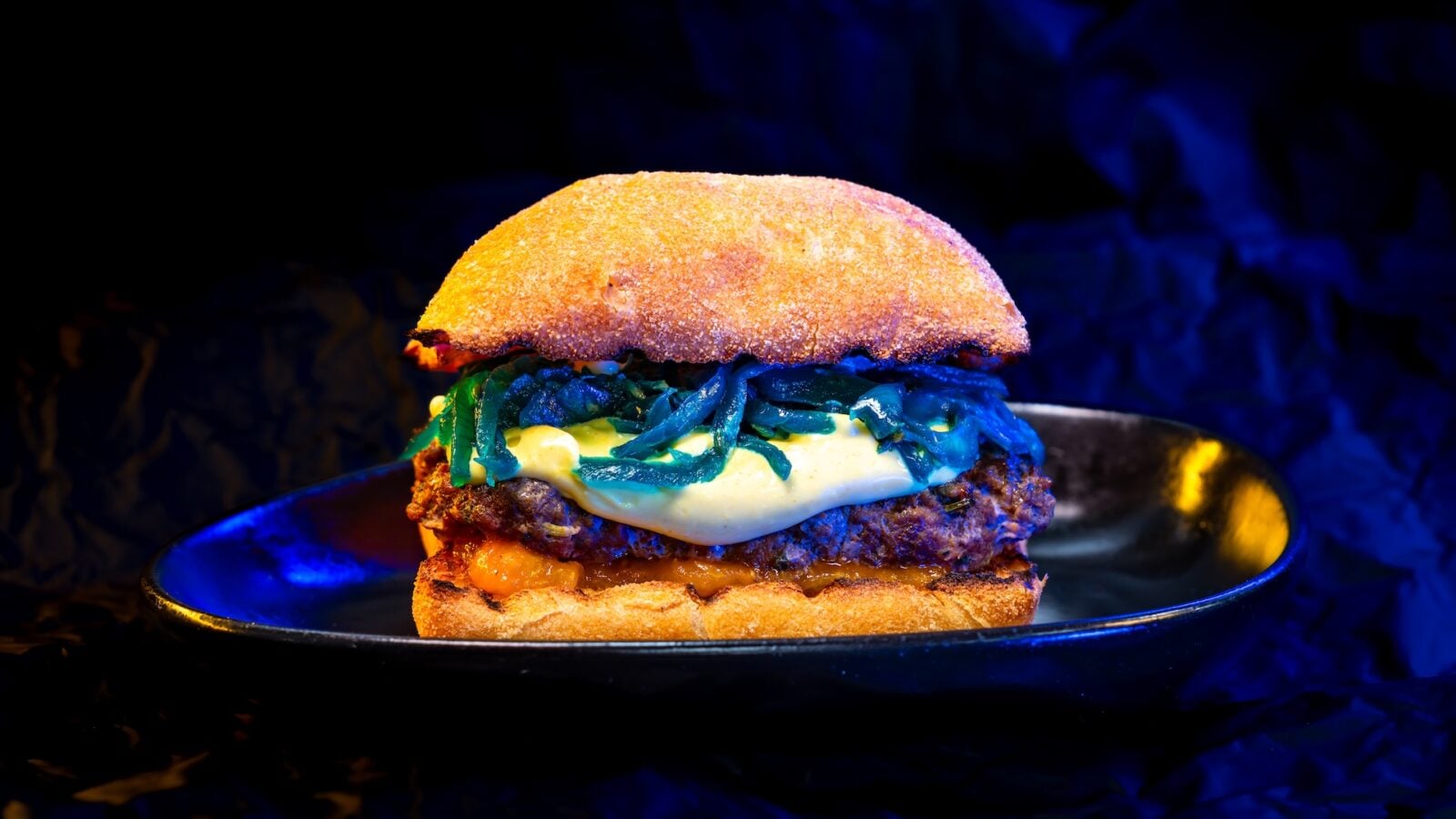 Close up of a cheeseburger with melted cheese and sautéed greens between toasted buns on a dark plate under blue lighting