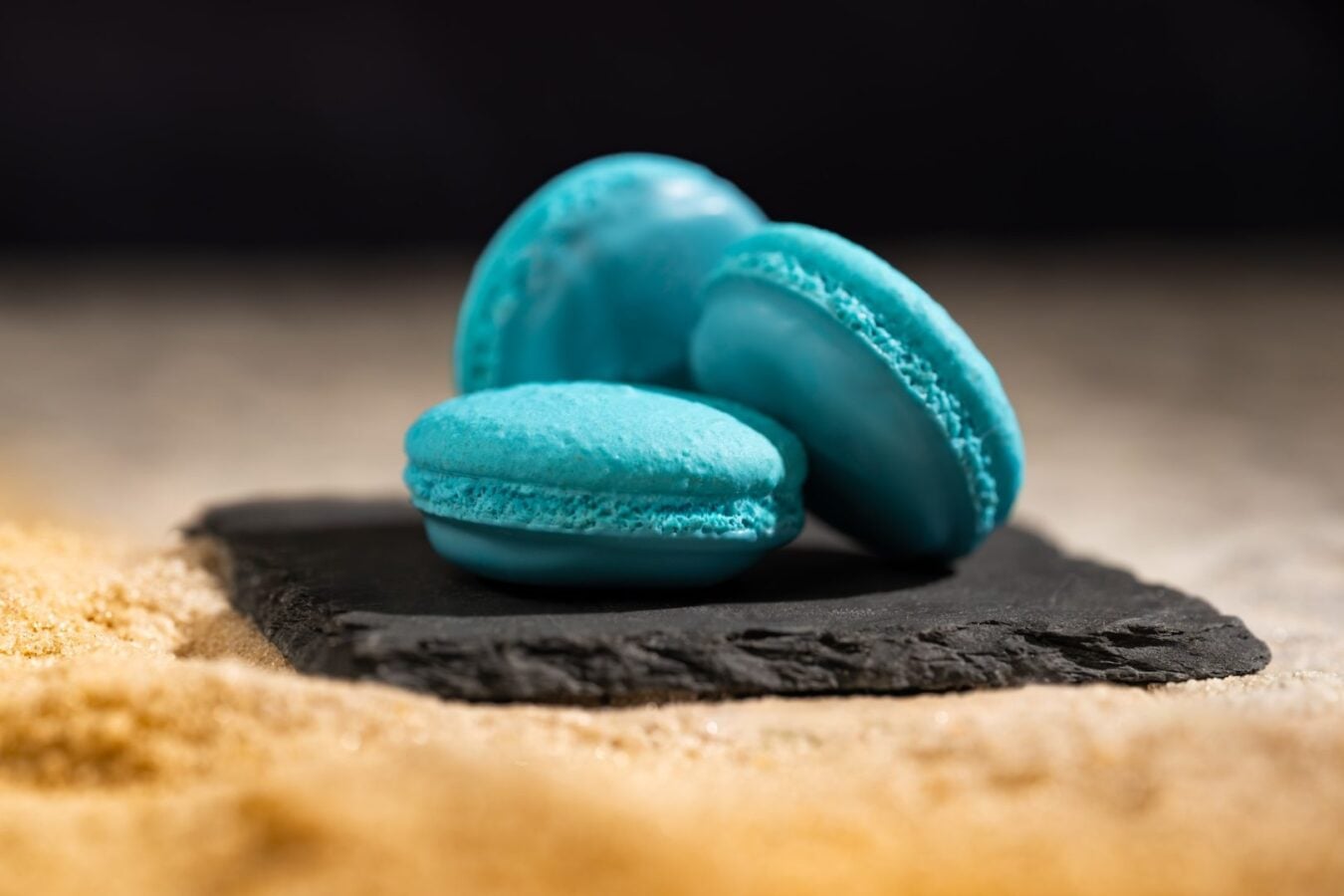 Blue macarons stacked on a dark slate board with sandy foreground