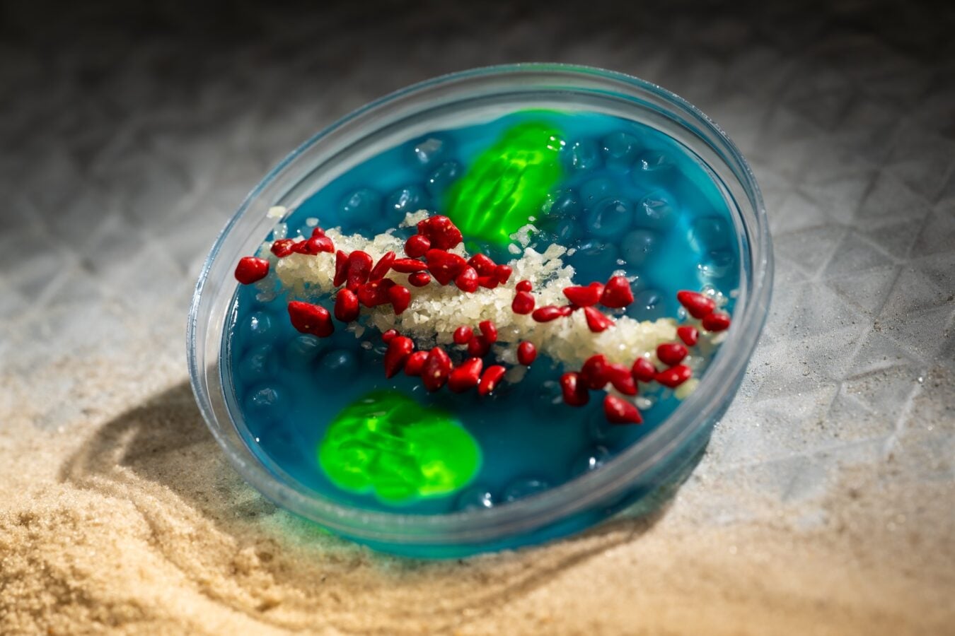 Close up of a clear petri dish with bright blue liquid red bead like pieces and white crystals resting on sandy surface