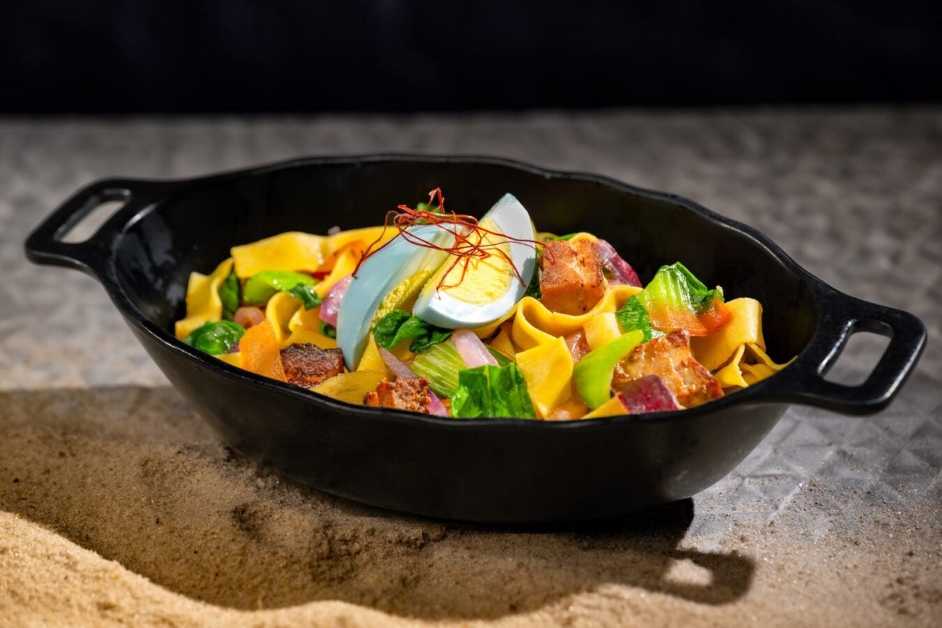 Pasta salad in a black skillet topped with a halved boiled egg and red chili threads served on a sandy surface