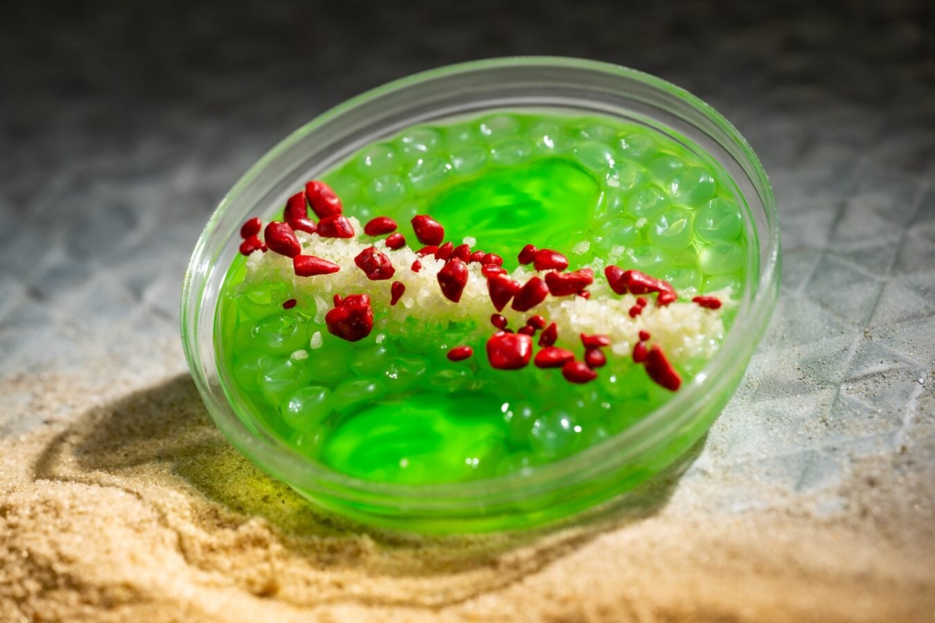 Close up of neon green slime in a clear container topped with red small chunks and white crystals set on sand