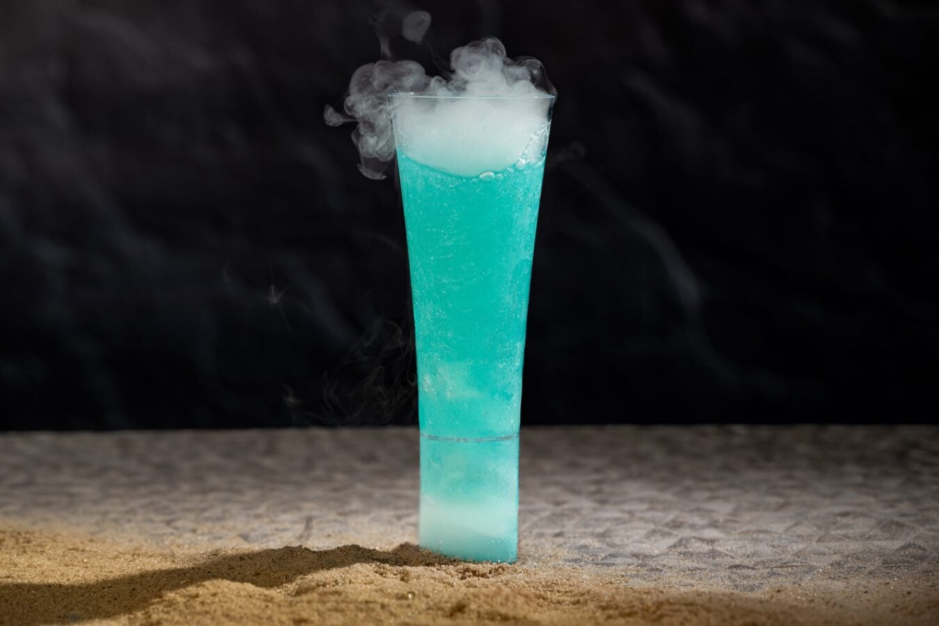 Tall turquoise cocktail in a narrow glass with fog from dry ice set on a sandy surface against a dark background