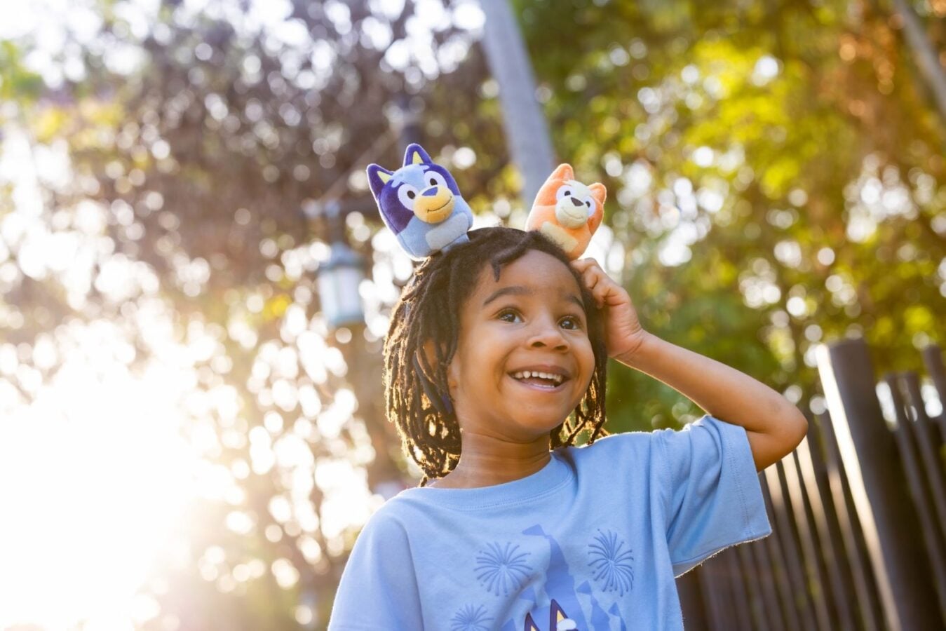 Smiling child in a blue shirt wearing stuffed animal ear headbands outdoors on a sunny day