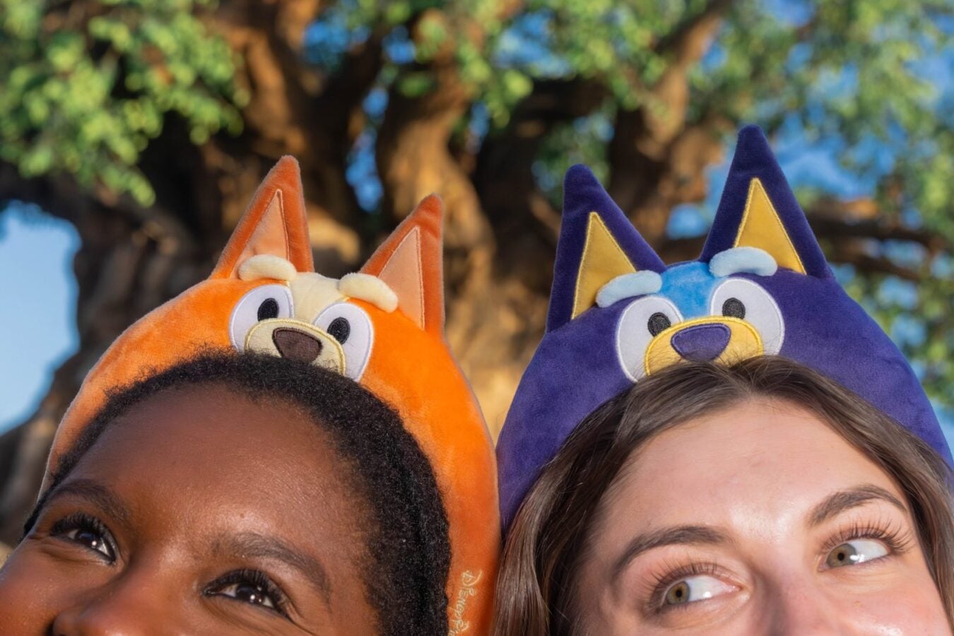 Two people wear bright animal hood hats orange fox and purple wolf and smile during an outdoor selfie with a tree in the background