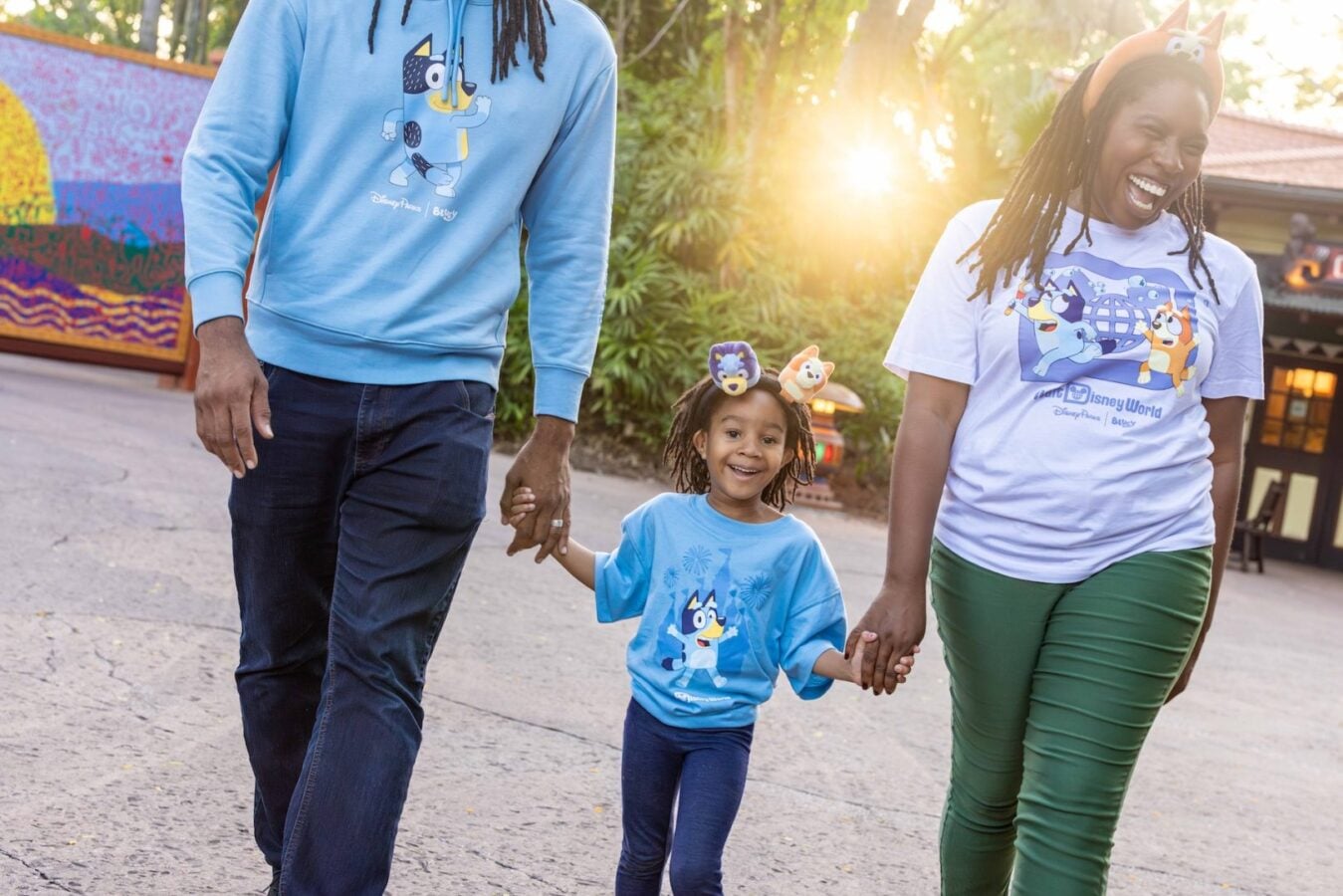 A joyful family of three walking hand in hand outdoors child in center wearing a blue shirt with a cartoon character adults on either side in matching Disney themed shirts