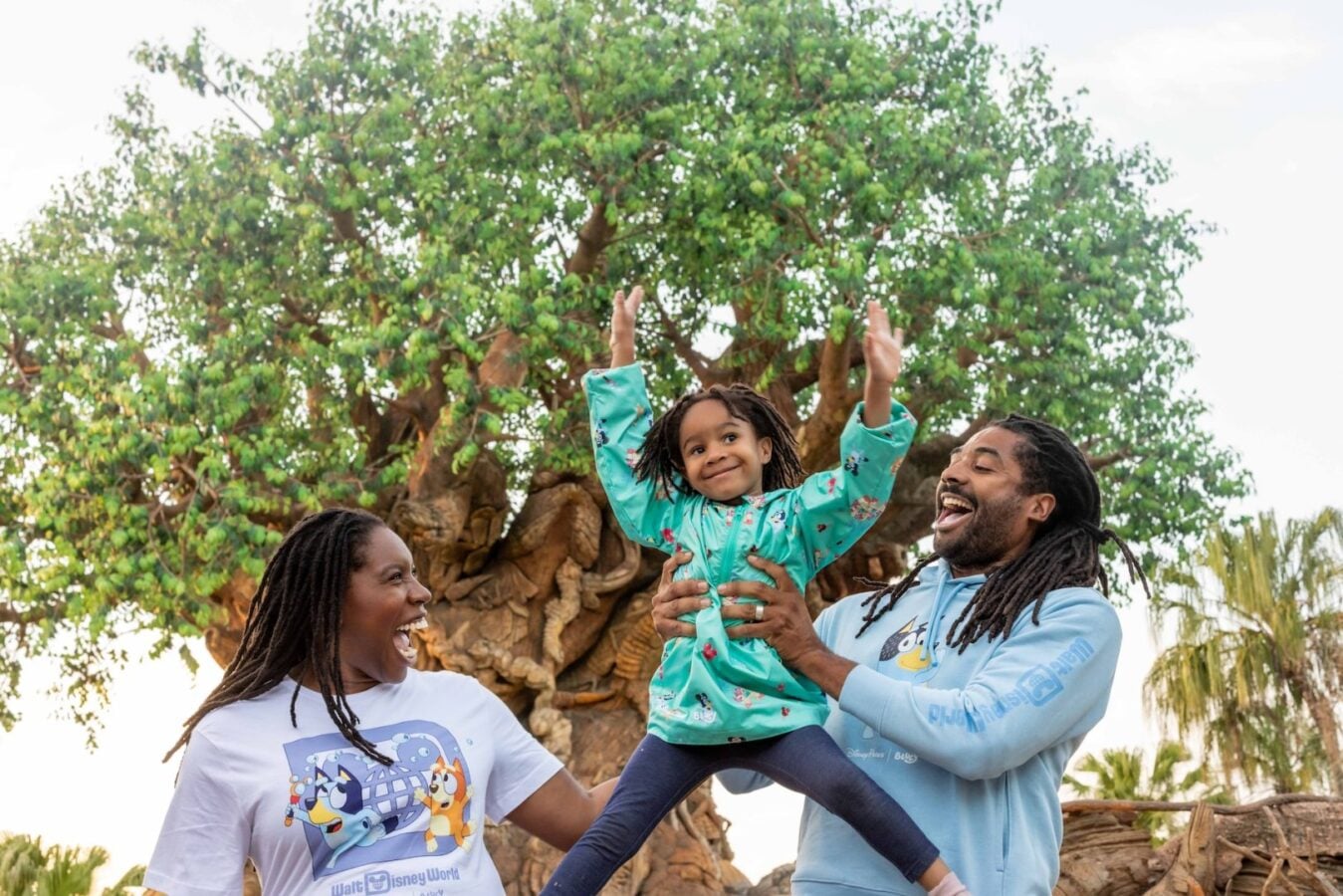 Parents lift their smiling child in the air outdoors all wearing matching Disney World shirts