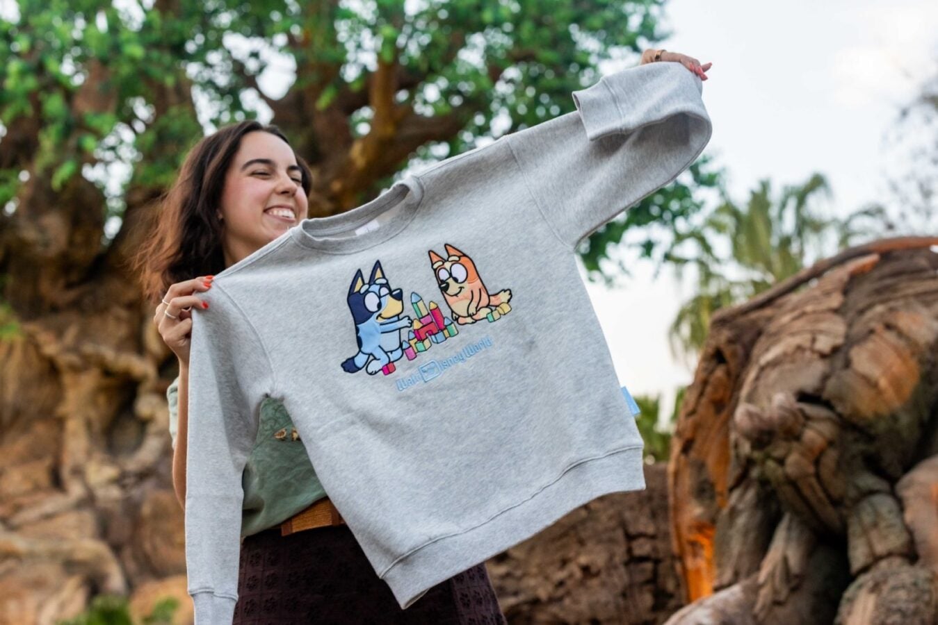 Woman outdoors holding up a gray sweatshirt with a cartoon blue and orange dogs building a block tower smiling face visible in background