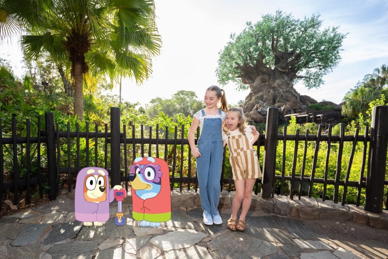 2026WDWBlueyAnimalKingdom81620x1080 Fantasy Land News Two smiling girls stand behind a black fence with cartoon character cutouts in front of them in a sunny garden