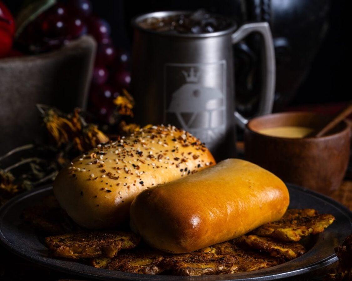 Two golden buns one sesame seeded sit on a dark plate with roasted potato wedges underneath and a dim kitchen backdrop