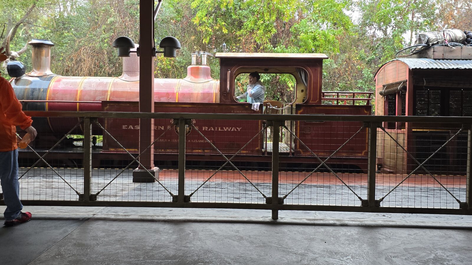 20260222100721 Fantasy Land News Red steam locomotive on a train platform with a metal railing in the foreground and a person in an orange jacket on the left