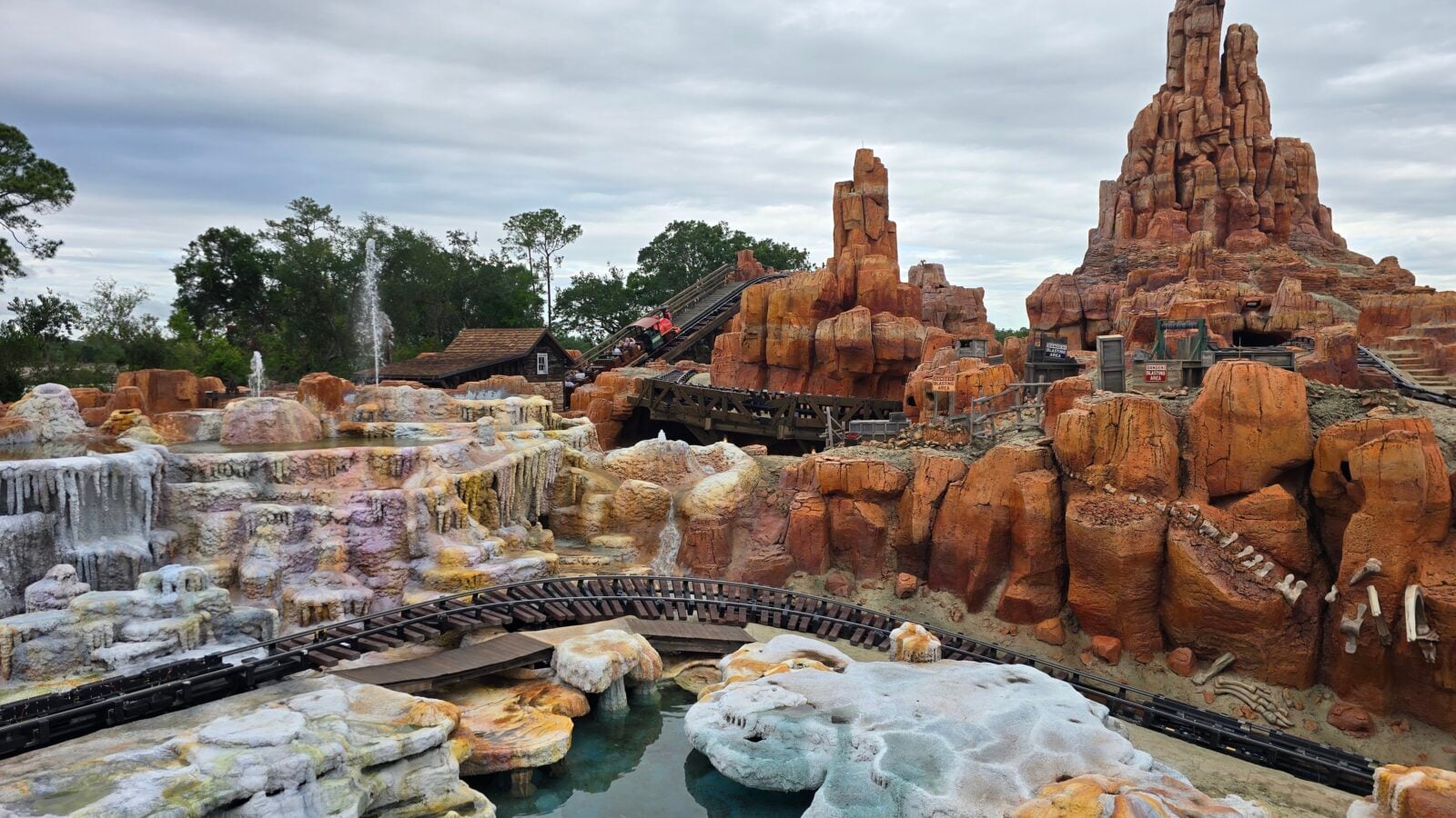 Orange red rock formations surround a wooden roller coaster track with a train of riders winding through the canyon a small pool in the foreground