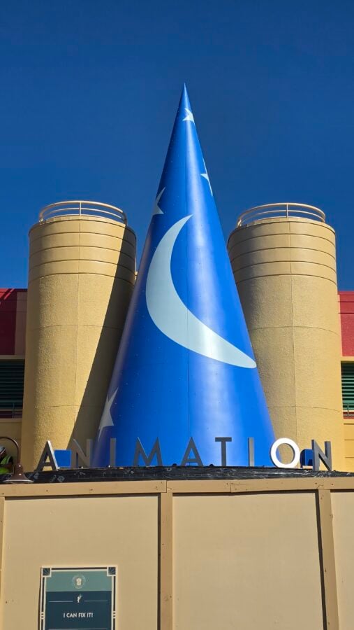 20260514095008 Fantasy Land News Tall blue cone sculpture with white stars in front of a beige building letters spelling ANIMATION visible below and a poster reading I CAN FIX IT