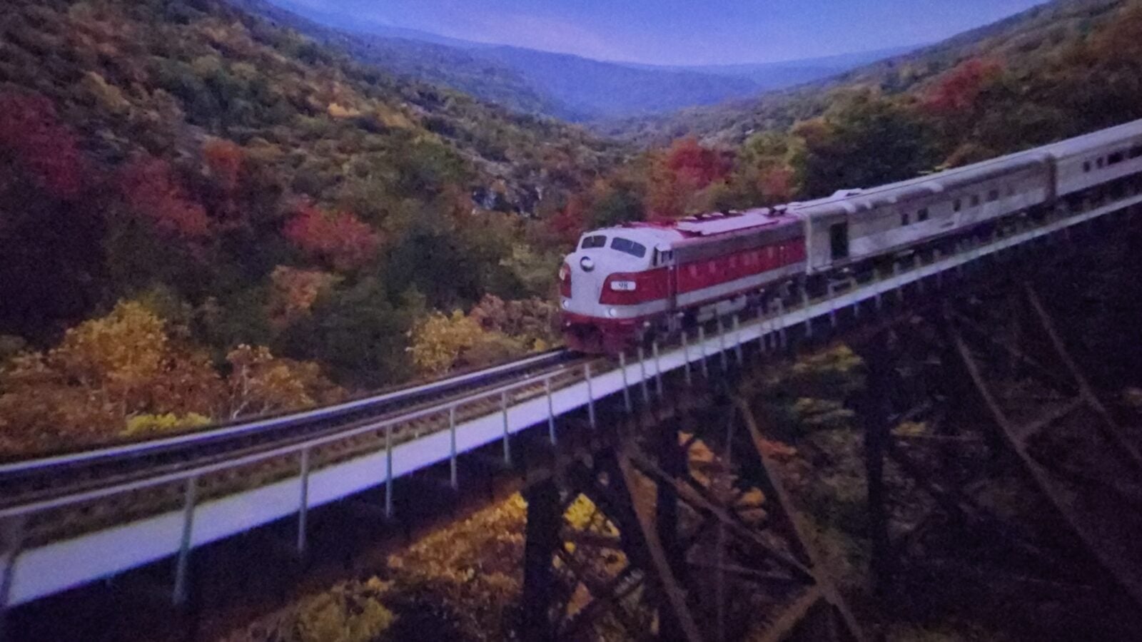 20260516102445 Fantasy Land News Red and white passenger train travels along an elevated track through a forest with vibrant autumn foliage at dusk