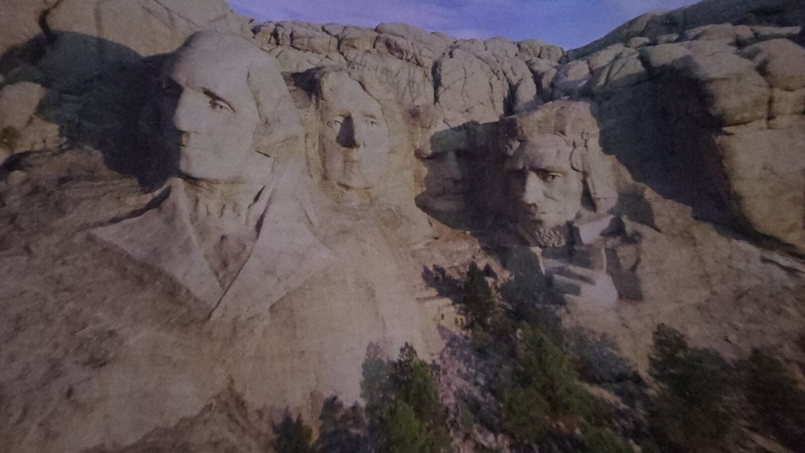 20260516102502 Fantasy Land News Rock carved mountain sculpture featuring four faces embedded in a rugged cliff with a purple evening sky and trees at the base nighttime view