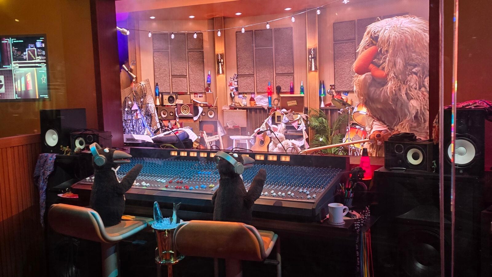 View into a recording studio through a glass wall a large shaggy puppet on the right and two plush figures with headphones at a big mixing console in the foreground
