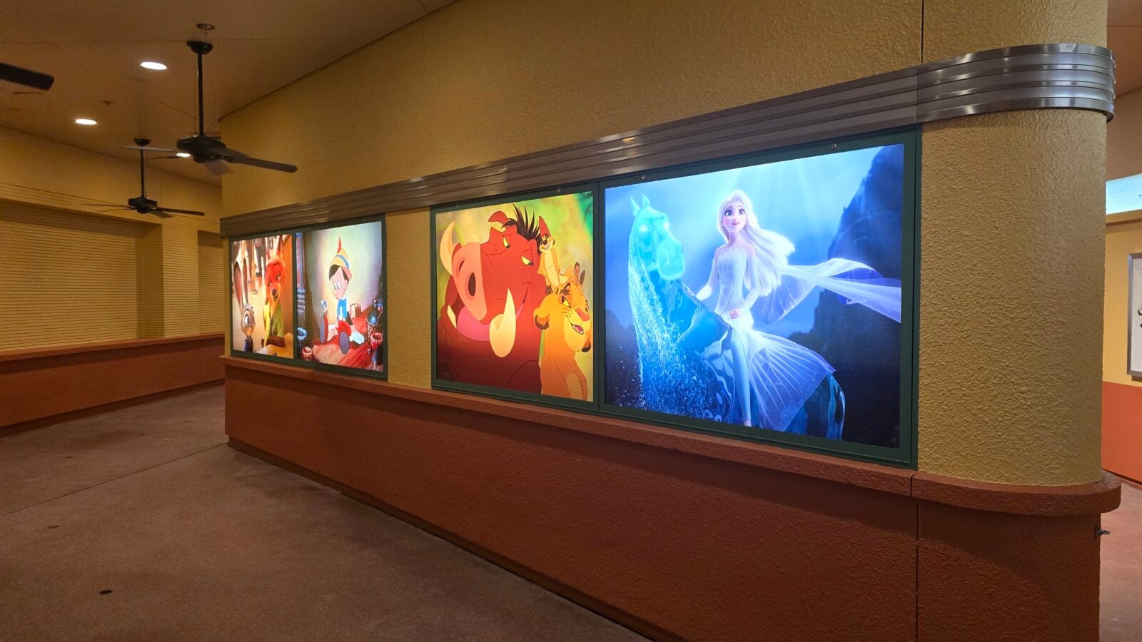 Row of four large animated posters mounted on a curved beige wall in a dim hallway including a blue Elsa from Frozen on the rightmost panel