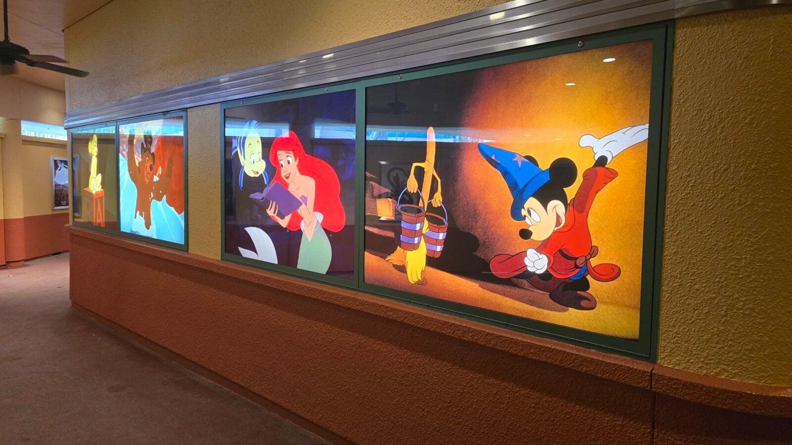 Hallway display with large glass panels showing Disney characters like Ariel reading a book and Mickey Mouse with buckets on an orange background