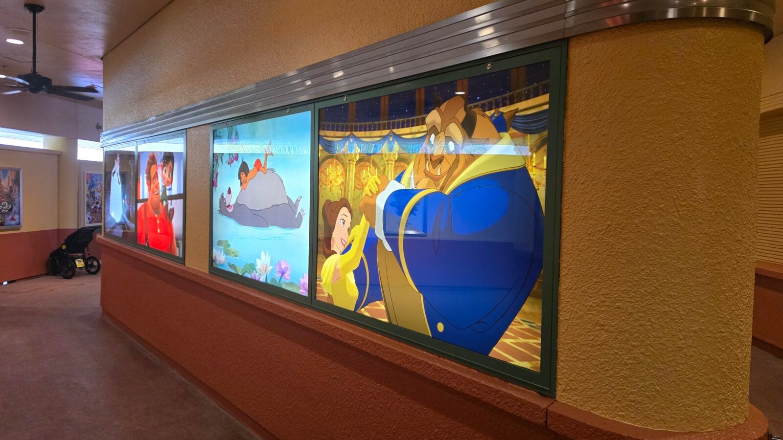 Indoor corridor with large Disney character posters in glass display cases along a textured brown wall features Belle and the Beast dancing in a ballroom scene on the right