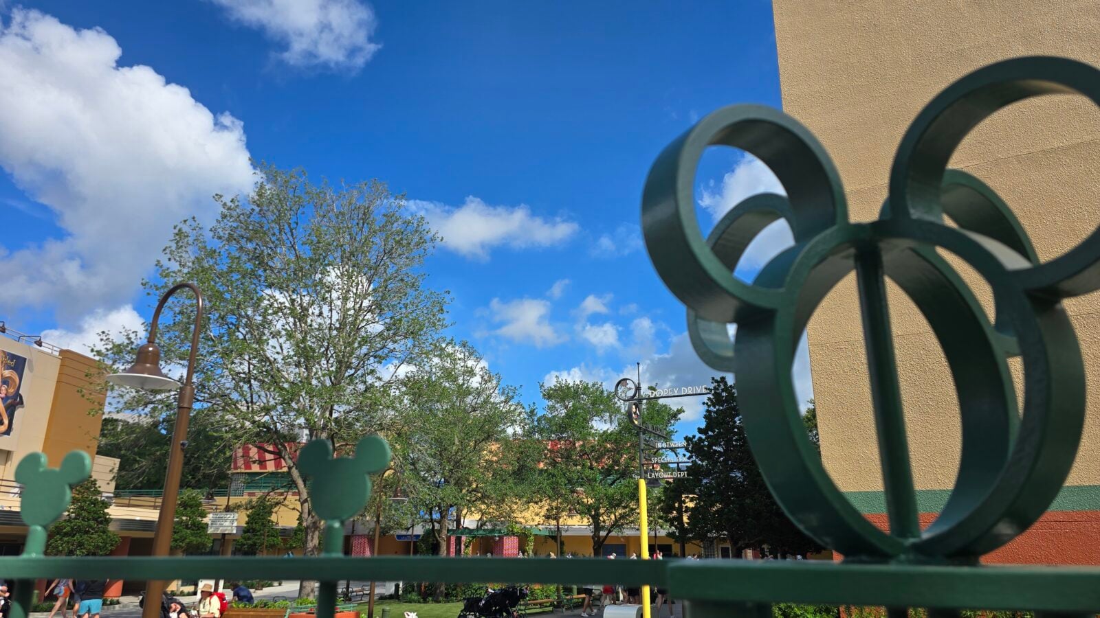 Blue sky with fluffy clouds above trees and a beige building seen through a dark green decorative metal sculpture in the foreground