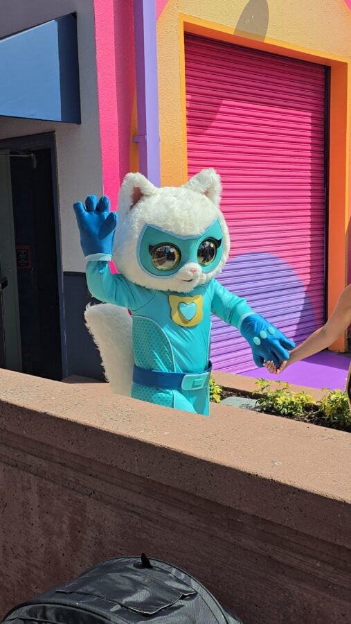 20260522104129 Fantasy Land News White fluffy cat mascot in a turquoise superhero suit waves hello while holding a childs hand against a vibrant pink purple and yellow mural backdrop