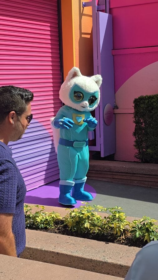 202605221041410 Fantasy Land News White cat mascot in a turquoise superhero outfit and blue boots stands outside a colorful pink and purple building greeting guests