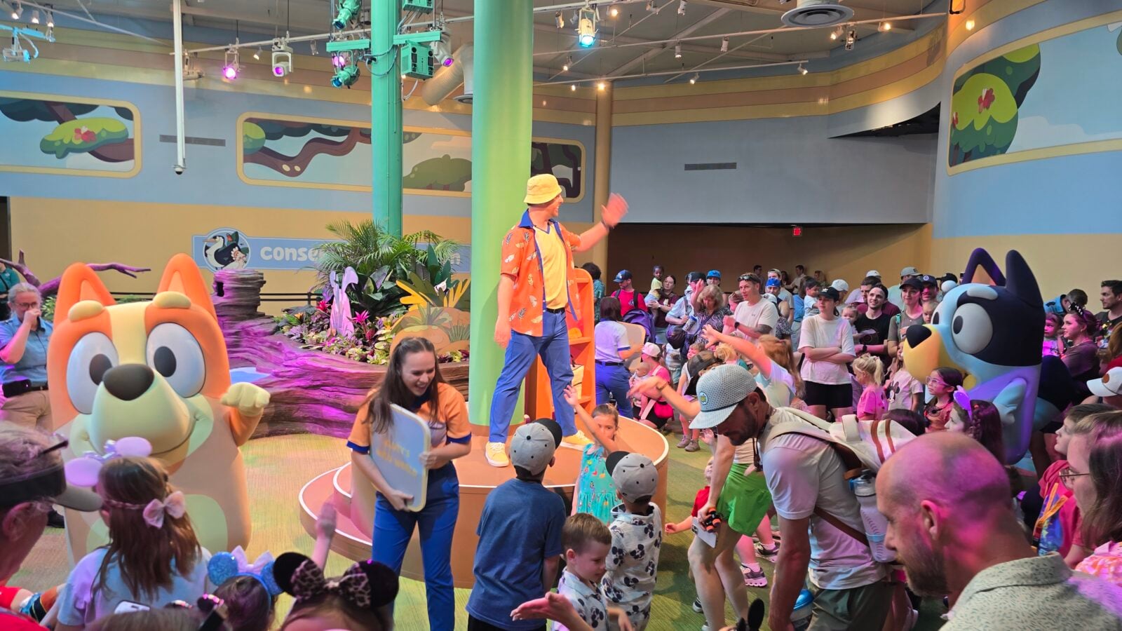20260525110000 Fantasy Land News Crowd of kids and adults watching a playful stage performance with colorful mascots and a performer in a bright outfit indoors