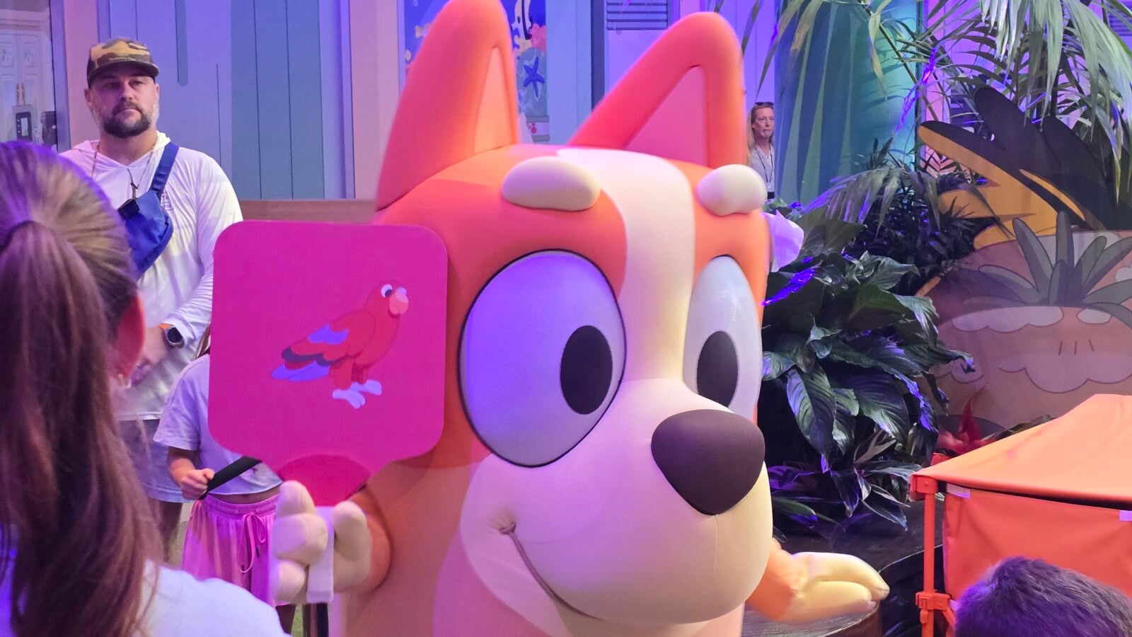 20260525110229 Fantasy Land News Large pink cartoon rabbit mascot with orange ears and big eyes at an indoor plant filled event with people nearby