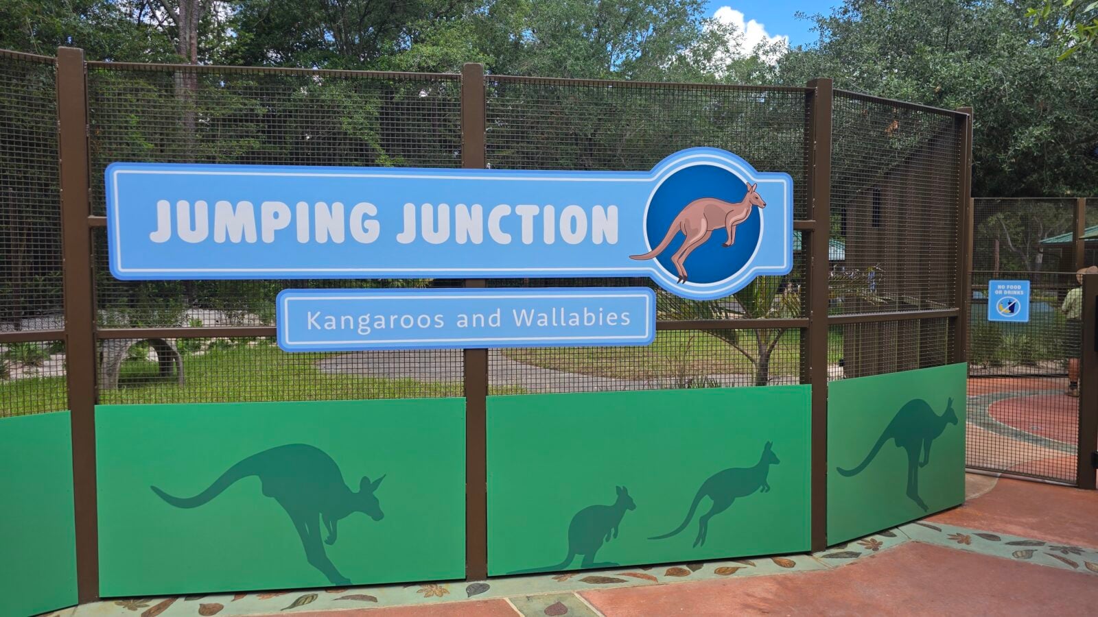 20260525112439 Fantasy Land News Sign reads Jumping Junction Kangaroos and Wallabies with a jumping kangaroo logo on blue panel behind a brown fence area