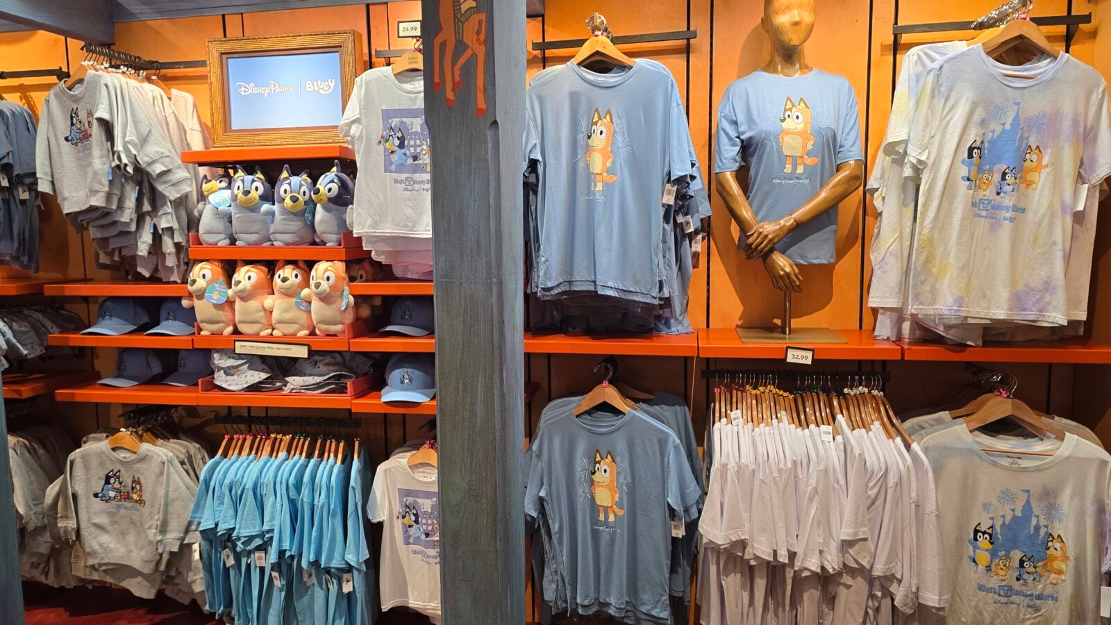 20260525123524 Fantasy Land News Display of blue and gray Disney World shirts with cartoon dog character and seasonal prints on orange shelves plus plush toys nearby