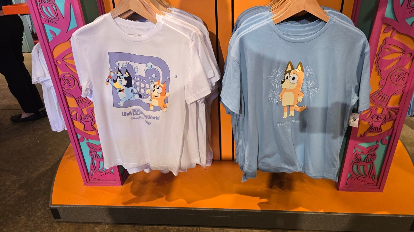 20260525123614 Fantasy Land News Two graphic t shirts on wooden hangers in a bright display a white tee with cartoon dogs in a spaceglobe scene and a light blue tee with an orange dog under fireworks on an orange pedestal