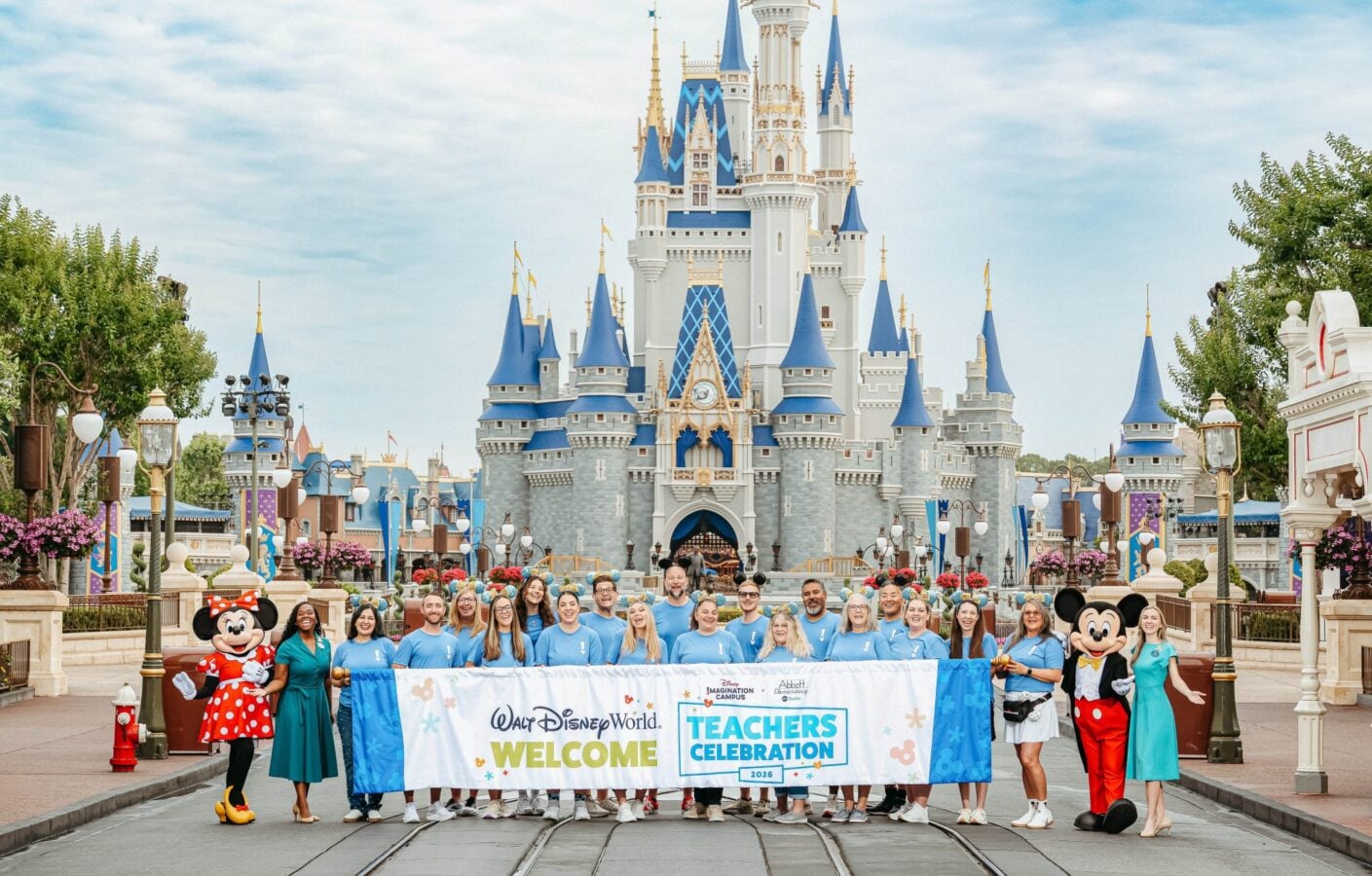 Group of teachers in blue shirts pose with Mickey and Minnie in front of Cinderella Castle at Walt Disney World for a Teachers Celebration banner and welcome event is shown in the foreground