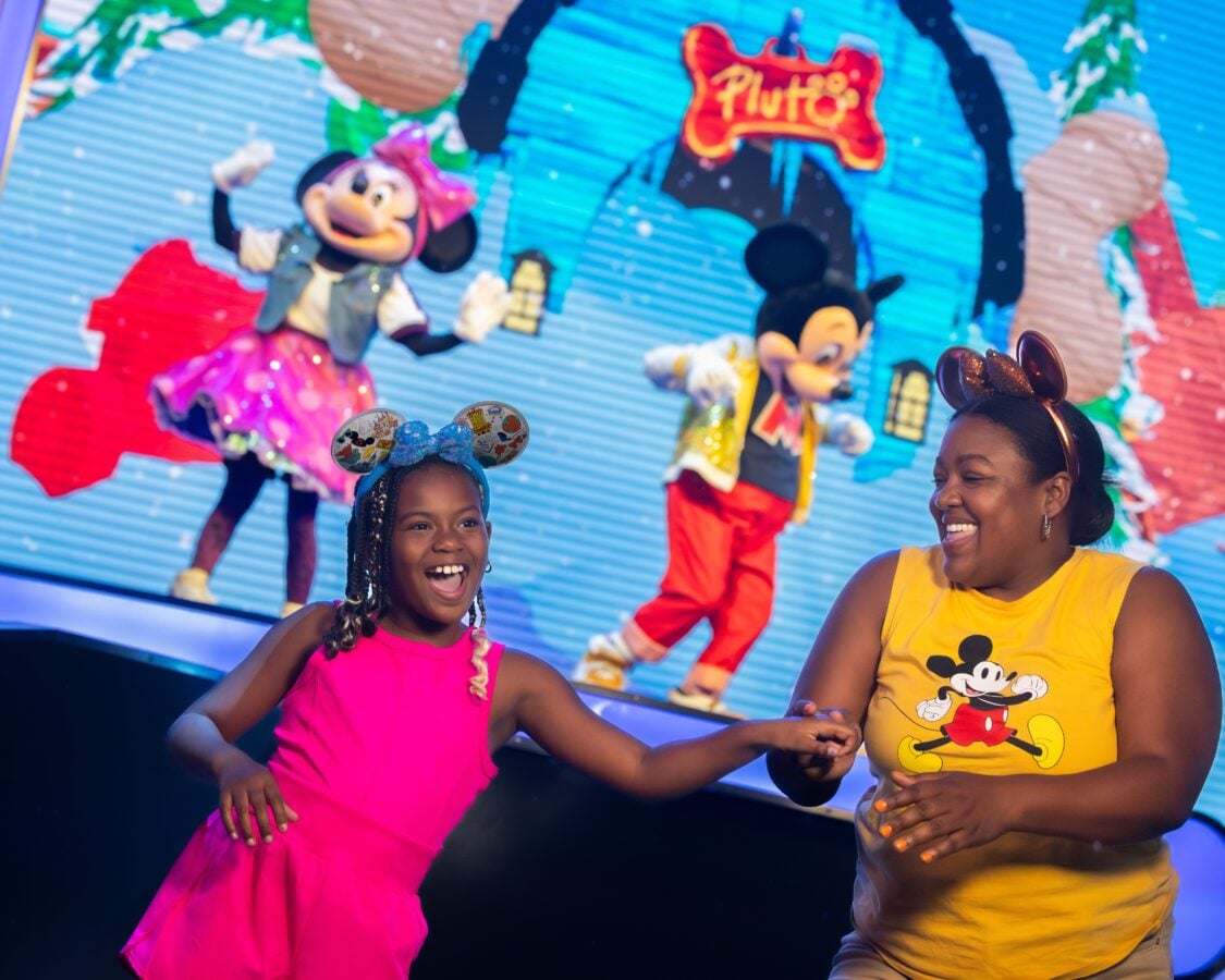Two smiling performers wearing MickeyMinnie ear headbands dancing on a colorful stage with a Disney themed backdrop