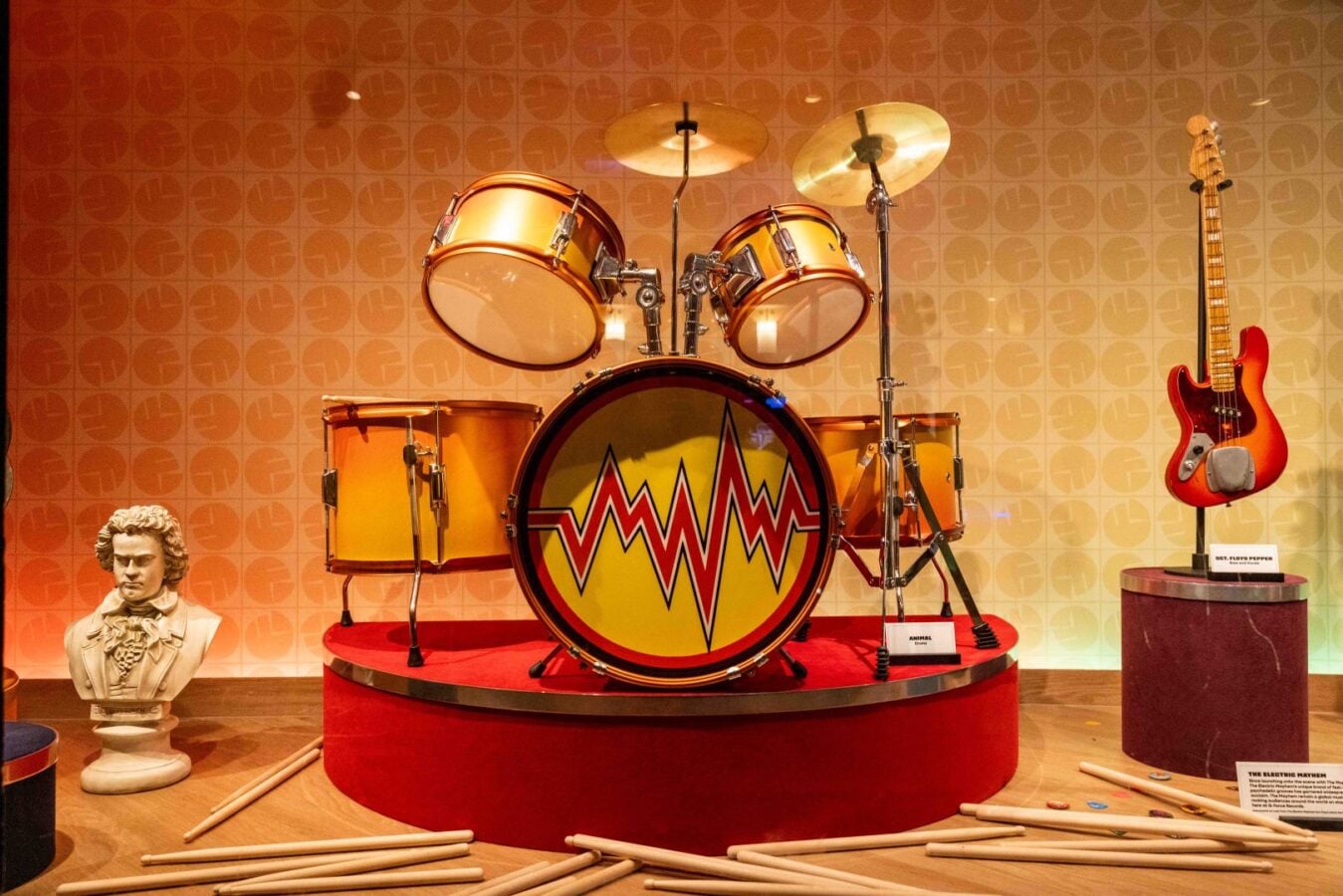 A vintage drum kit on a red pedestal in a museum display with a yellow zigzag pattern bass drum cymbals and an orange drum set accompanied by a red electric guitar on the right and a bust on the left