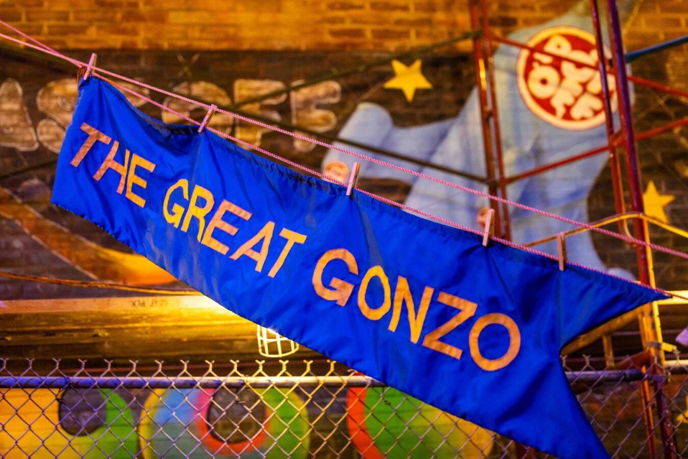 Blue banner reading THE GREAT GONZO hung on a clothesline with clothespins over a colorful mural and chain link fence in warm lighting
