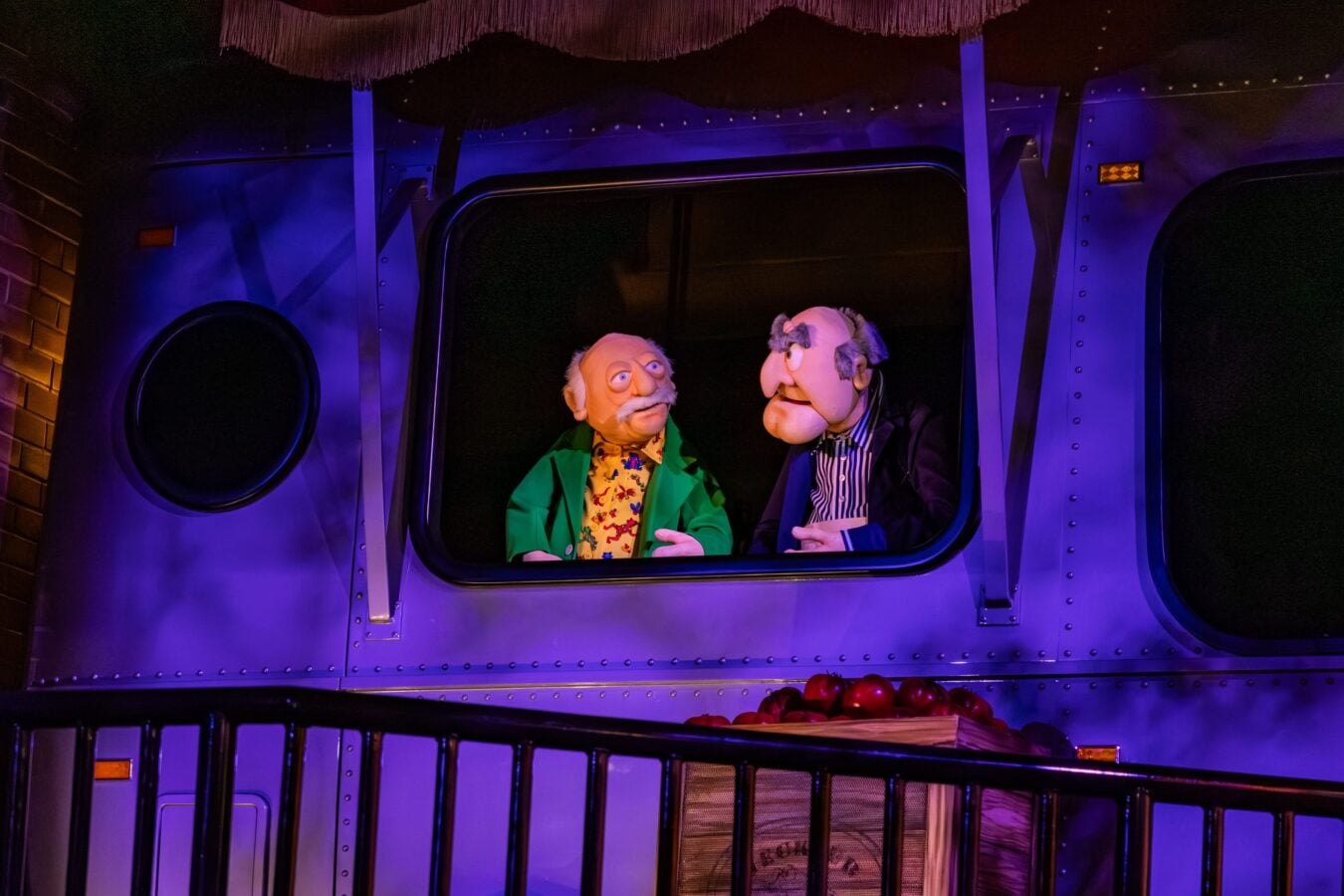 Two puppets peek through a bus window under blue purple lighting as if performing a show inside a vehicle