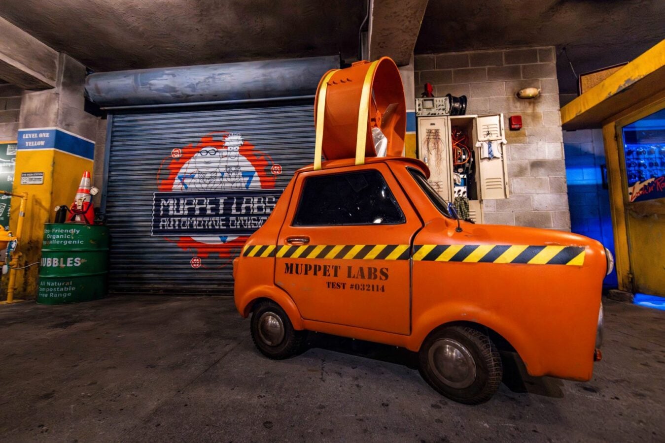 Orange compact car labeled Muppet Labs with black and yellow caution stripes parked inside a workshop with a large orange helmet like device on its roof