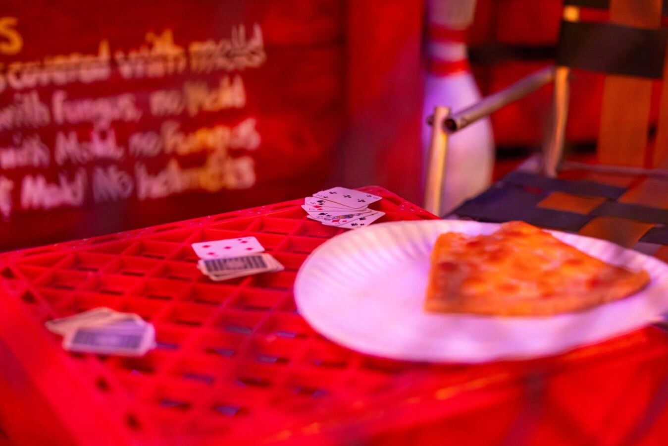 A slice of pizza on a white plate with a red plastic table and scattered playing cards in a dim colorful bar setting