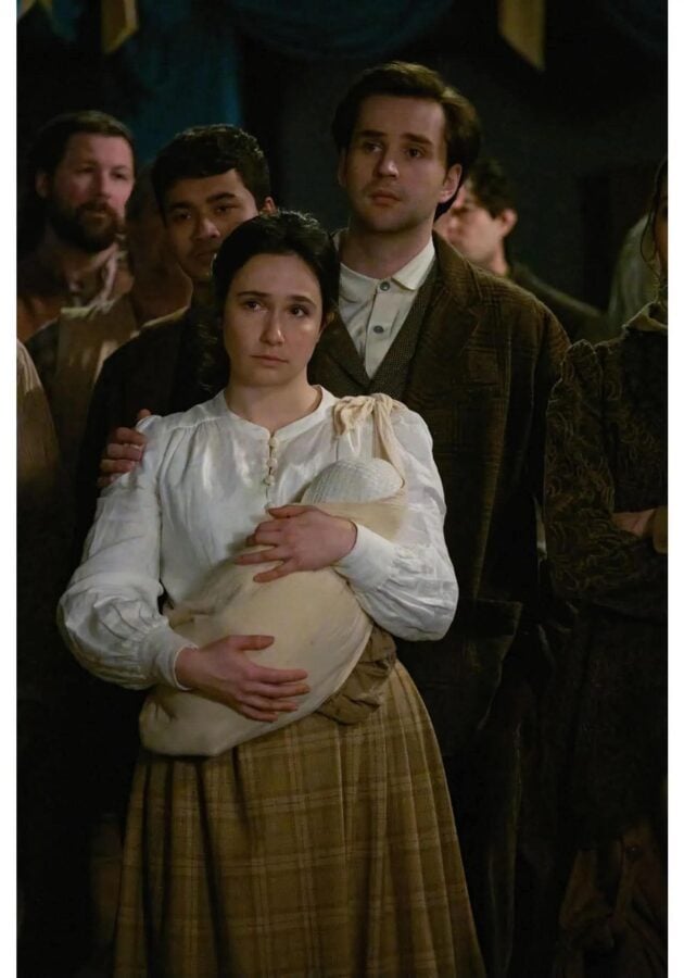 Woman in a white blouse cradles a wrapped baby while a man in a plaid coat stands close behind in a dimly lit crowd