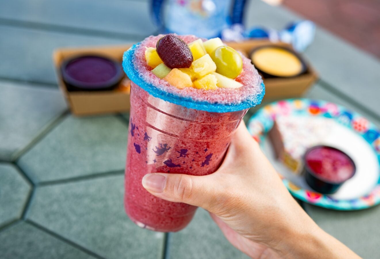 Pink shaved ice beverage with a blue sugar rim topped with mixed fruit being held outdoors by a hand