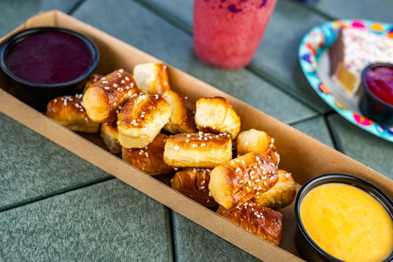 Soft pretzel bites with coarse salt in a cardboard box paired with a yellow mustard dip in a cup at a picnic table