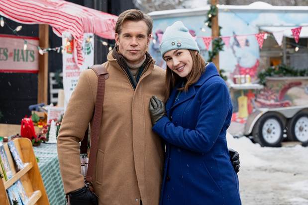6a19bca0325e4 Fantasy Land News A couple posing together in a snowy outdoor market man in a tan coat and scarf woman in a blue coat and light blue beanie smiling at the camera