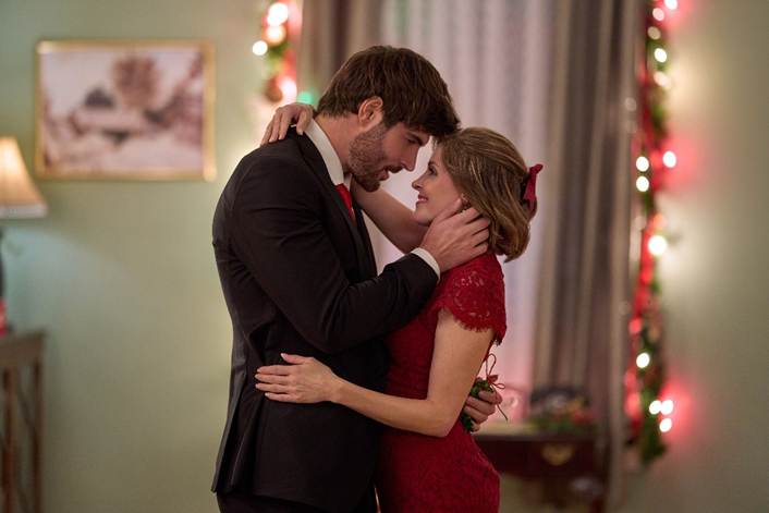 6a19bca0d17a4 Fantasy Land News Couple sharing a kiss while embracing in a festively lit room with red dress and holiday lights in the background