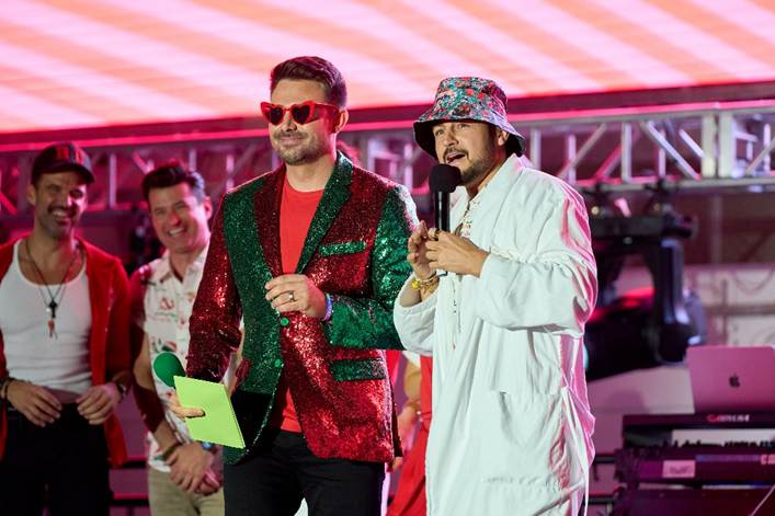 6a19bca110437 Fantasy Land News Two male hosts on a stage with a microphone and clipboard one wears a shiny green red sequin blazer and red sunglasses the other a white bucket hat and light jacket as a band watches from behind