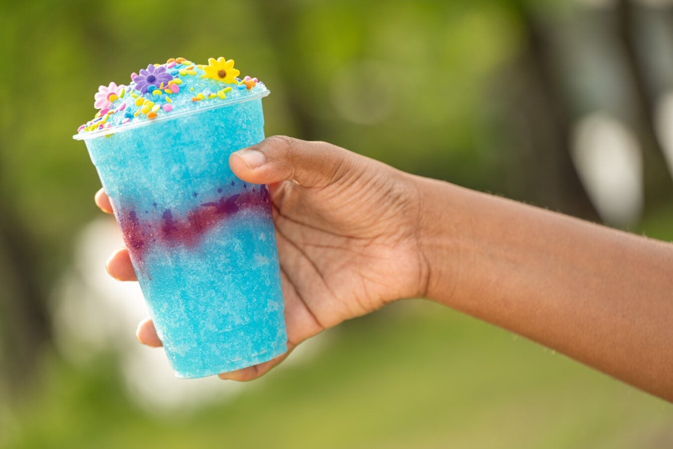 Hand holding a blue gradient frozen drink topped with colorful sprinkles and candy flowers outdoors
