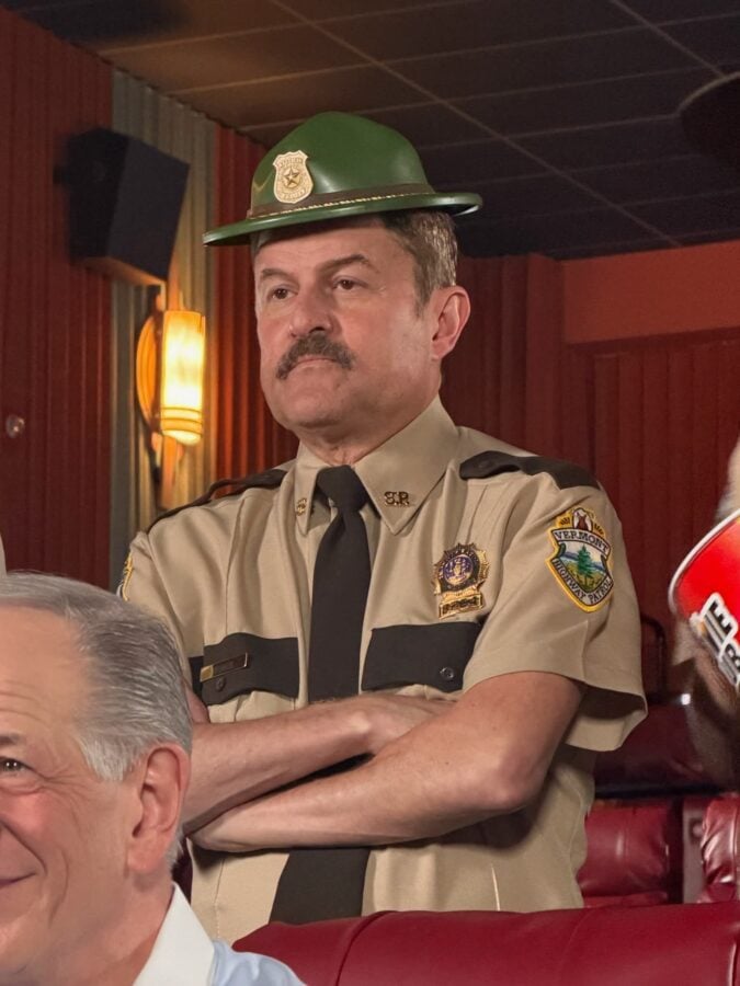 Male sheriff in beige uniform with badges and a green campaign hat standing with arms crossed in a wood paneled room