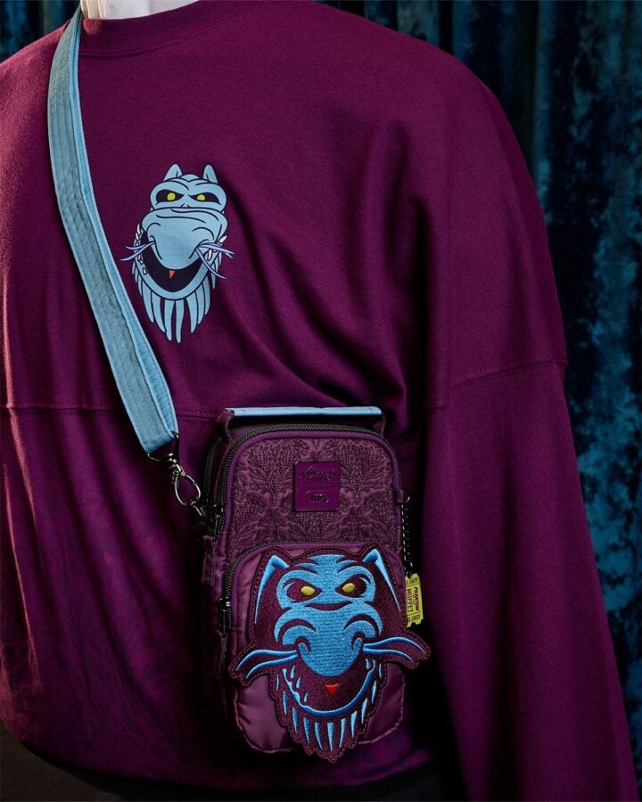 Close up of a purple sweatshirt with a blue cartoon monster logo on the chest and a small purple crossbody bag with a blue monster face hanging from the strap