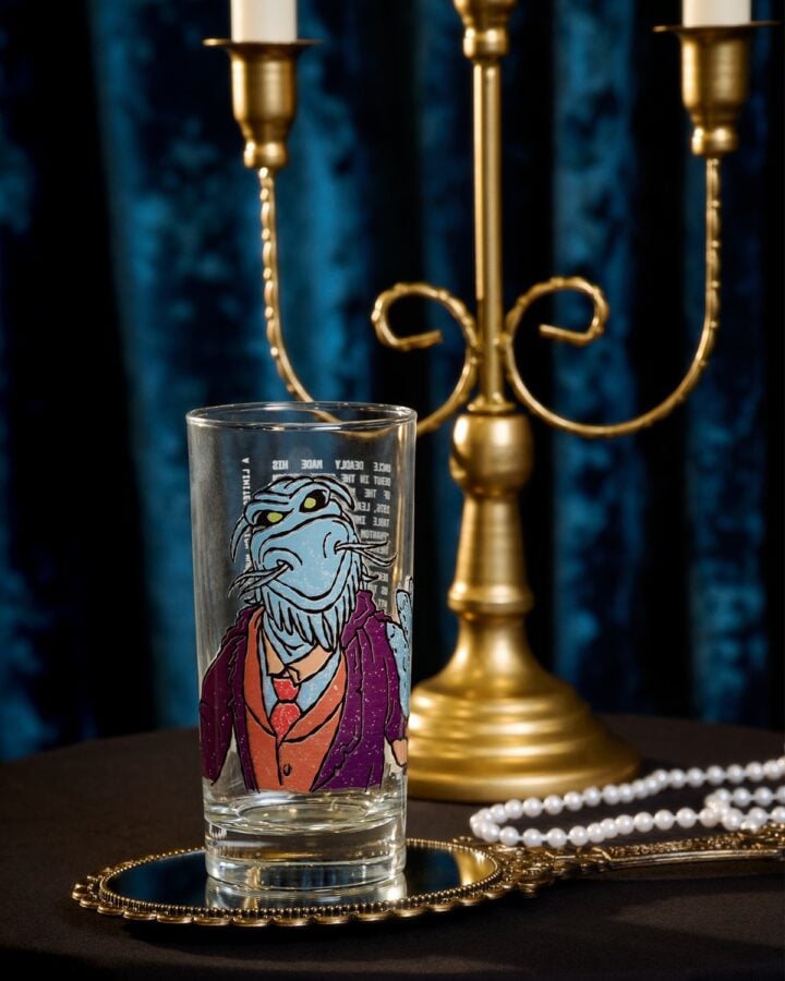 Glass featuring a blue cartoon alien in a purple jacket and red tie perched on a gold decorative tray with a pearl necklace and a brass candelabrum in the background
