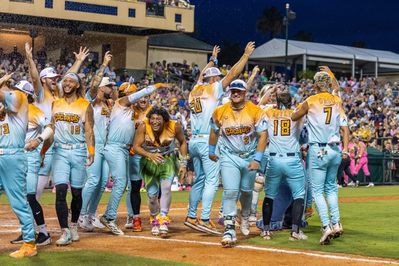 Team of softball players in teal and orange uniforms celebrate together on the infield as fans cheer in the stands at night
