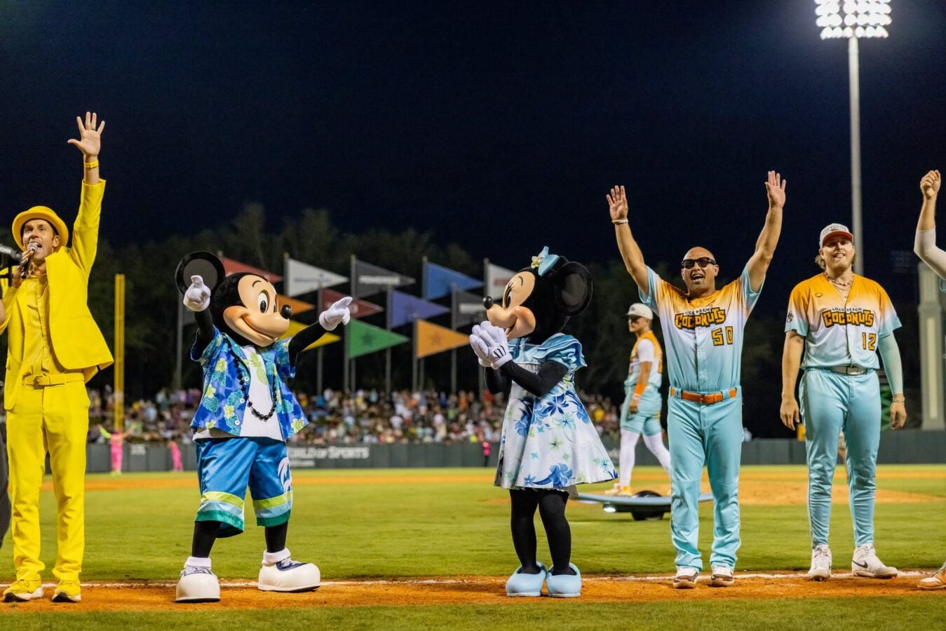 Night game on a baseball field with Mickey and Minnie Mouse mascots in Hawaiian shirts and players in teal uniforms waving to the crowd under bright stadium lights