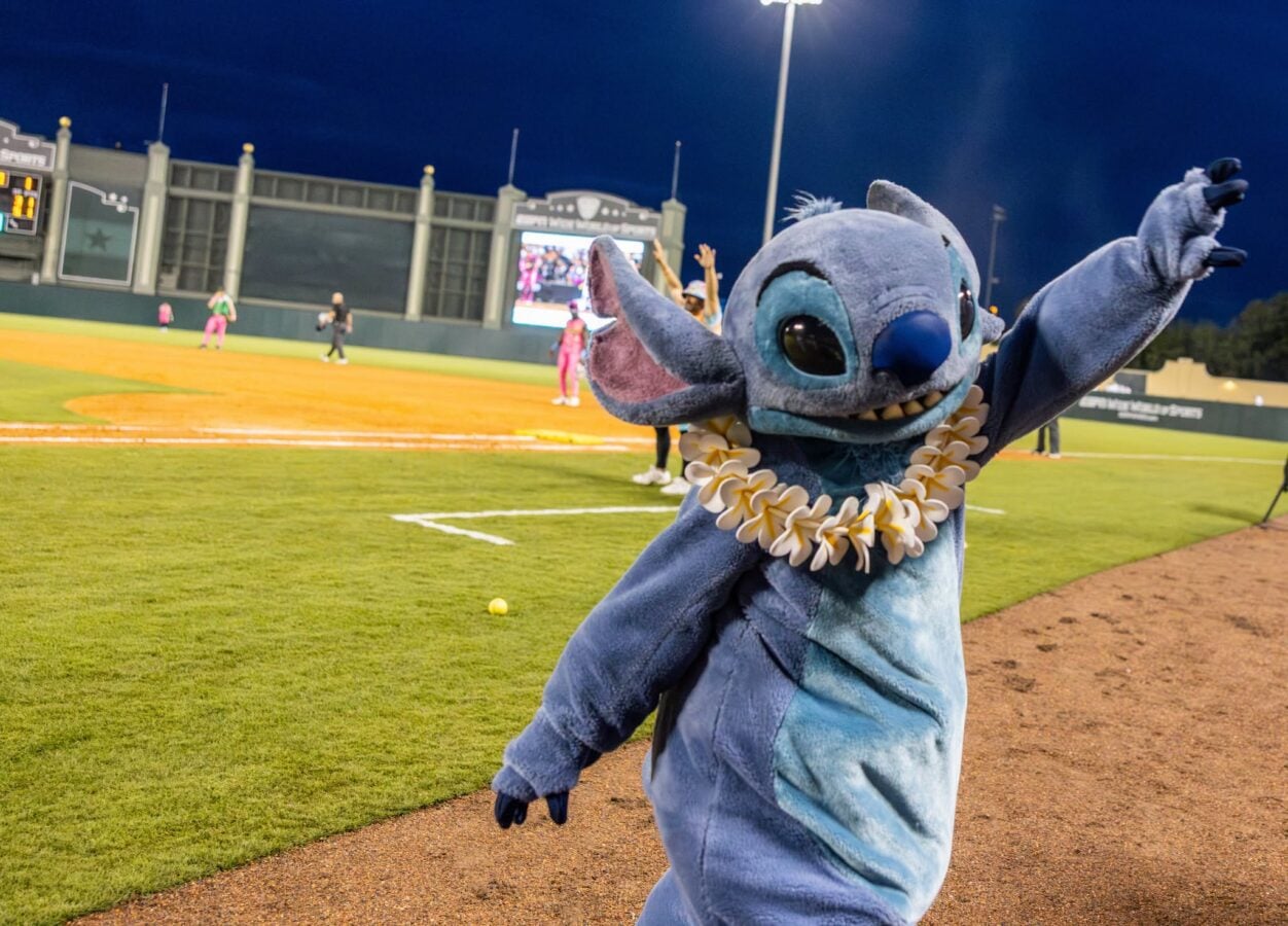 Stitch mascot in a blue costume with a flower lei arms raised on a baseball field at night